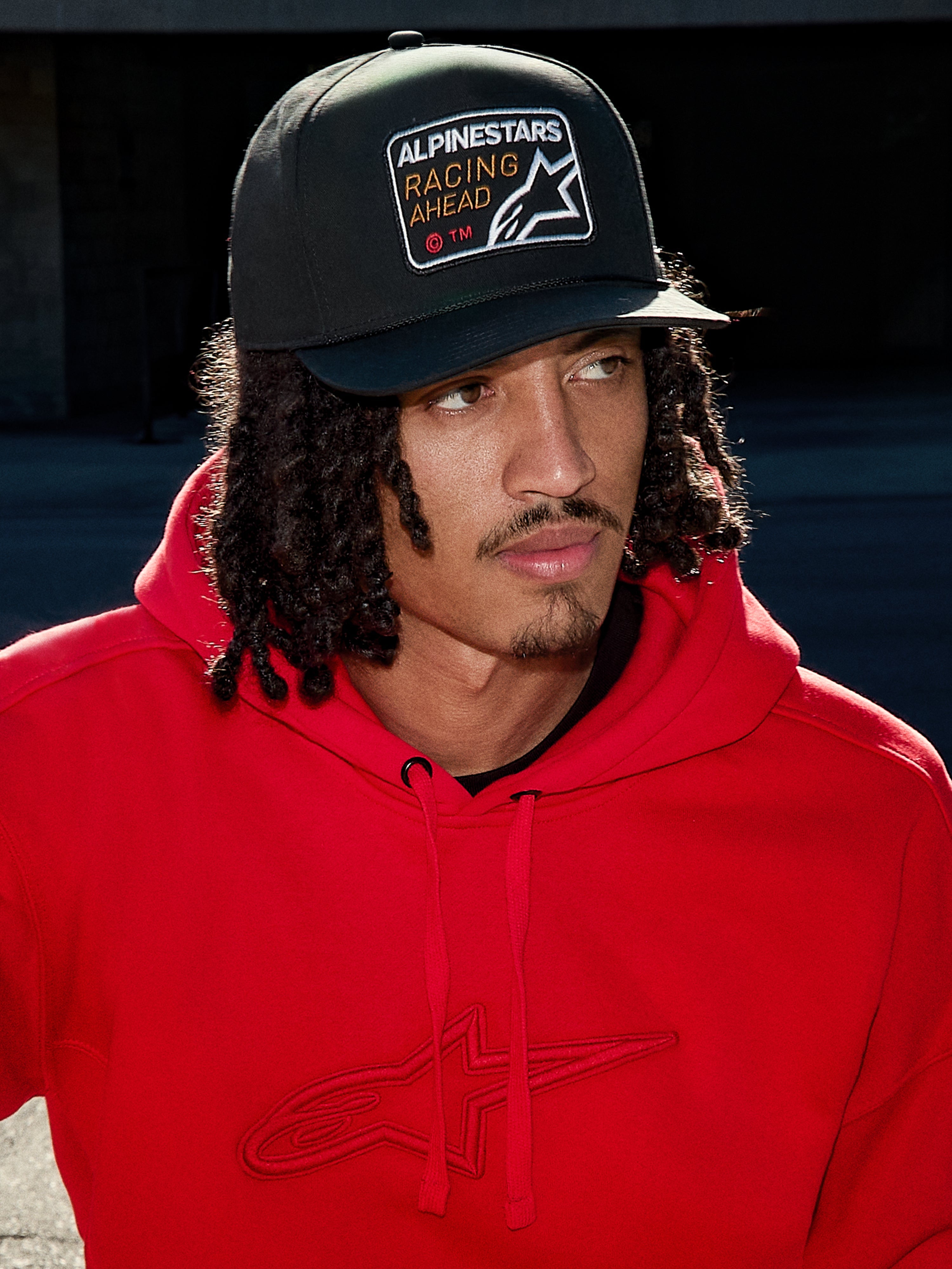 Hombre con la Gorra Snapback Intuitive de Alpinestars en color negro, con corona estructurada y parche bordado con el logotipo 'Racing Ahead', combinada con una sudadera roja brillante con capucha, estilo urbano casual
