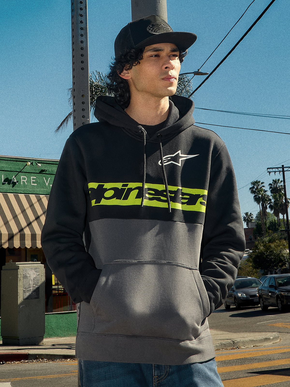 Sudadera con capucha Parallel de Alpinestars, estilo urbano lifestyle, diseño de bloques de color en negro y gris plomo, franja verde lima en el pecho con el logo de Alpinestars, logo Astars blanco, capucha con cordones, usada por un hombre en un entorno urbano al aire libre.