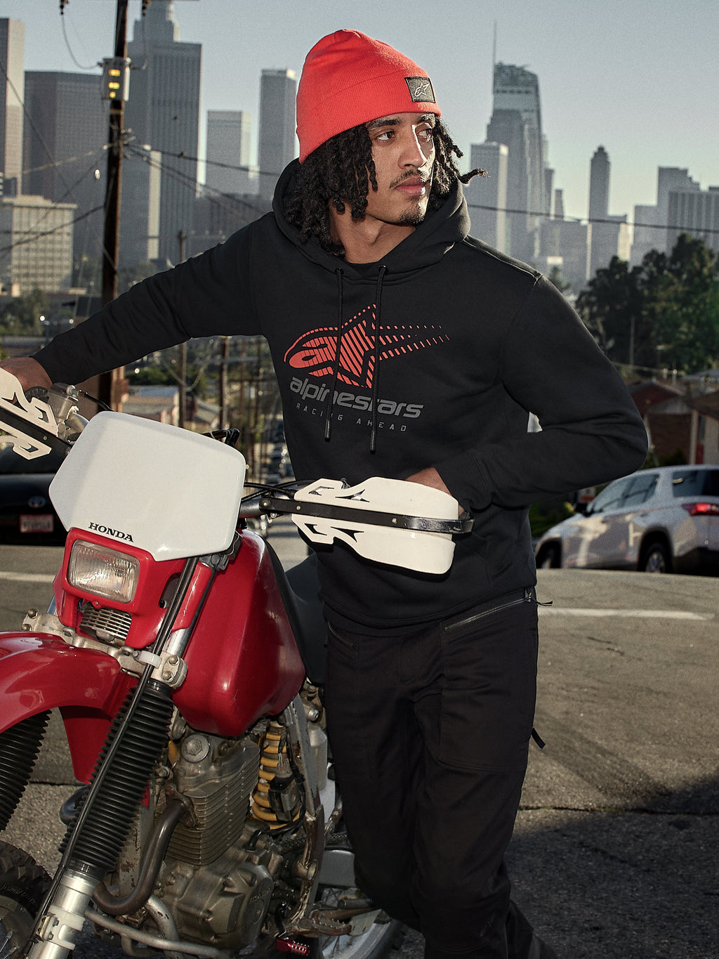 Sudadera con capucha Alpinestars Pulse, negra, sudadera de forro polar con logotipo Astars Combo en rojo y gris, hombre con gorro naranja junto a una motocicleta Honda roja en un entorno urbano, ropa urbana casual de estilo motorsport