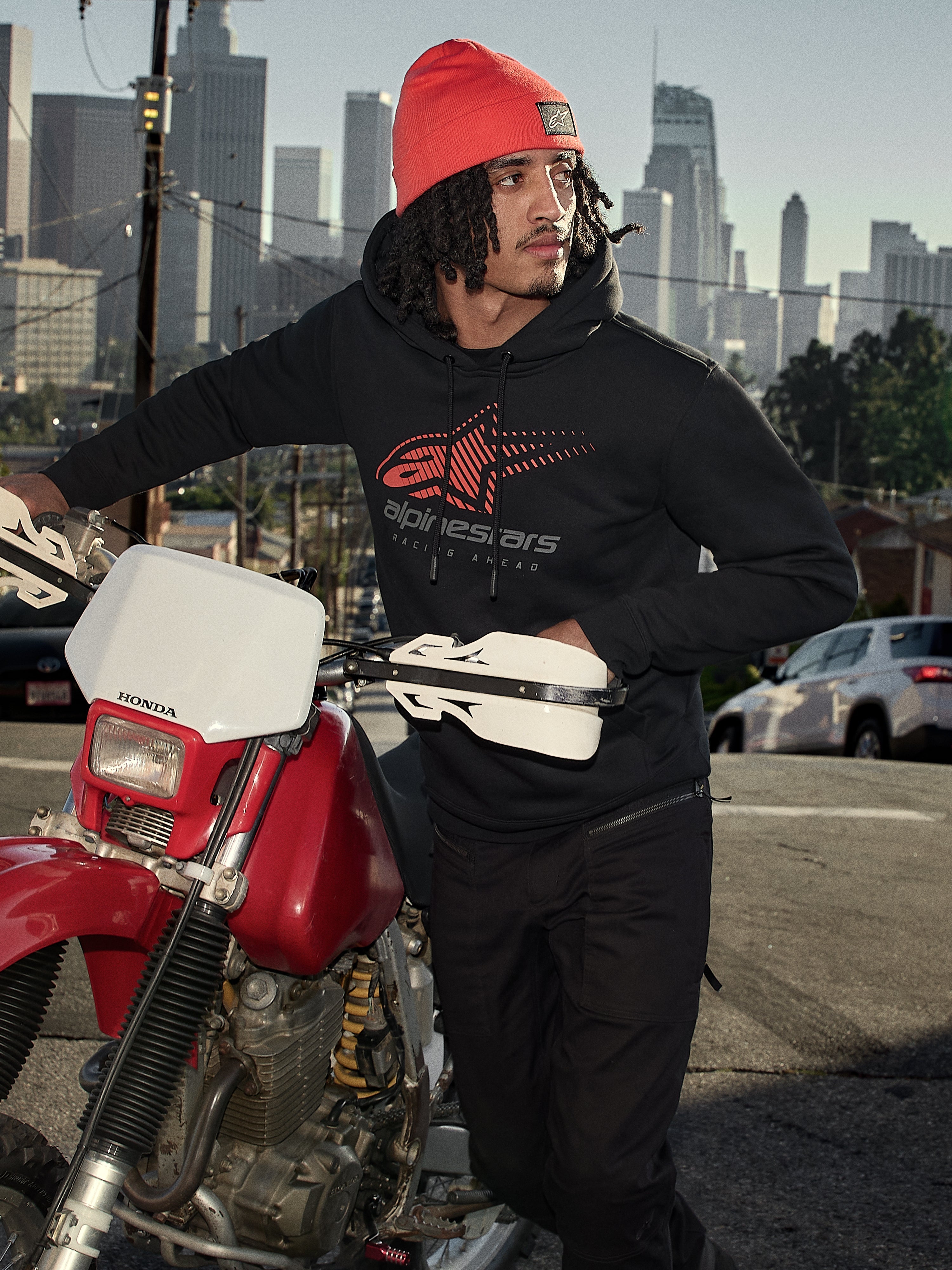 Sudadera con capucha Alpinestars Pulse, negra, sudadera de forro polar con logotipo Astars Combo en rojo y gris, hombre con gorro naranja junto a una motocicleta Honda roja en un entorno urbano, ropa urbana casual de estilo motorsport