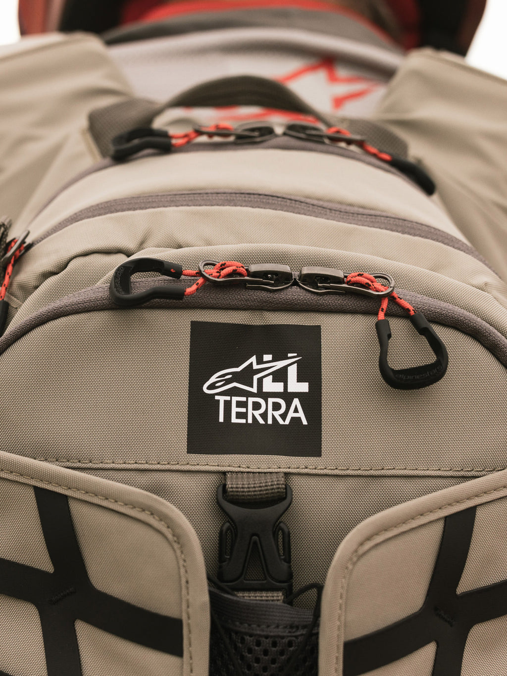Mochila Alpinestars AT-15, Aluminum Walnut, primer plano del parche con el logotipo All Terra sobre tejido técnico color arena, con tiradores de cremallera de cordón naranja y detalles de construcción robusta