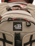 Mochila Alpinestars AT-15, Aluminum Walnut, primer plano del parche con el logotipo All Terra sobre tejido técnico color arena, con tiradores de cremallera de cordón naranja y detalles de construcción robusta