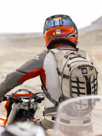 Mochila Alpinestars AT-15 en Aluminum Walnut, llevada por un motociclista de aventura en un paisaje desértico. Cuenta con una construcción textil beige resistente con correas negras, detalles naranjas en las cremalleras y el logotipo 'All Terra'. Combinada con un casco rojo y naranja y equipamiento gris para touring off-road.