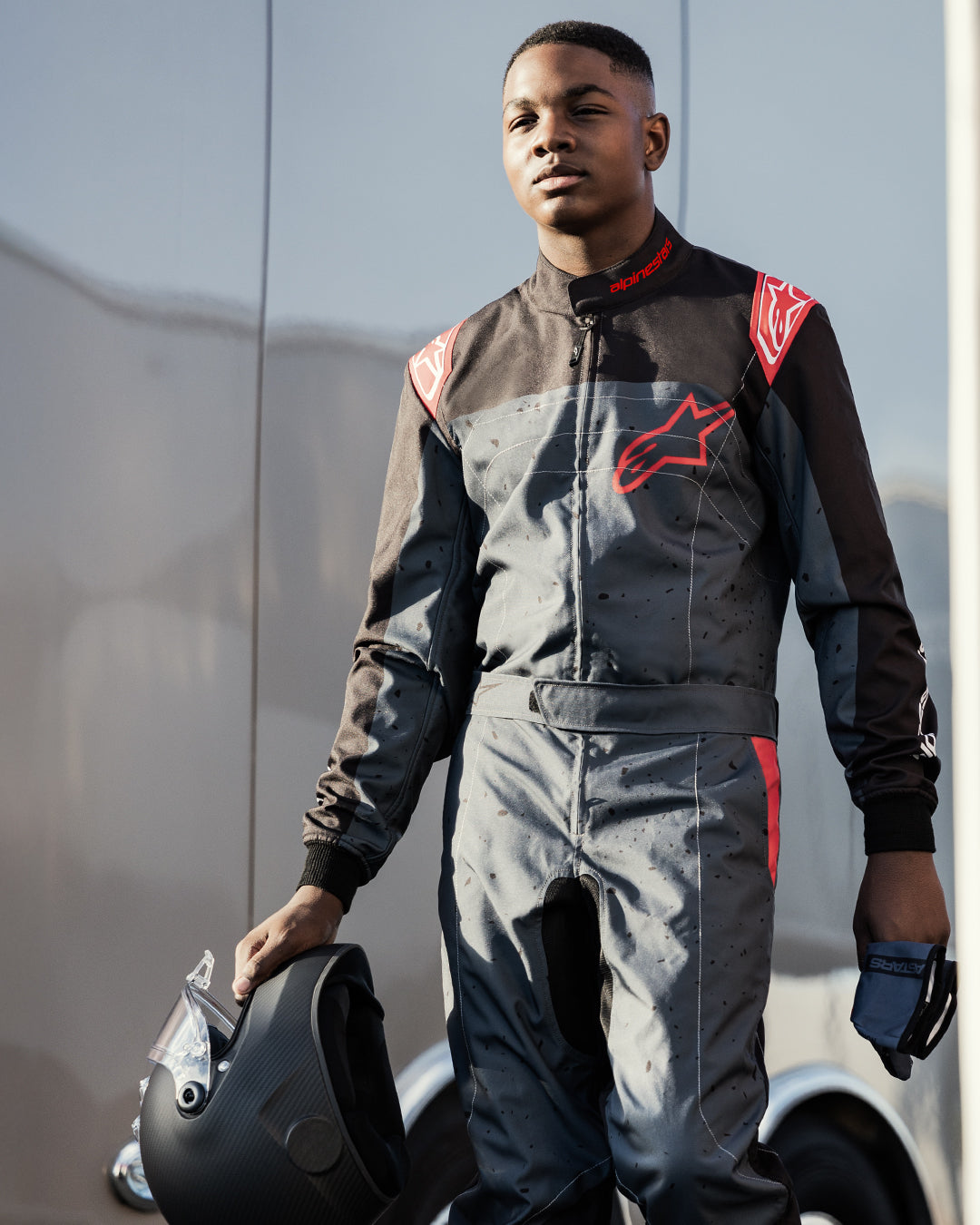 Piloto de karting con el mono Alpinestars KMX-9 V3 Graphic 6 en negro antracita y rojo, sosteniendo un casco negro mate y guantes, con logotipos rojos llamativos de Alpinestars y un diseño gráfico de salpicaduras, en un entorno de automovilismo al aire libre.