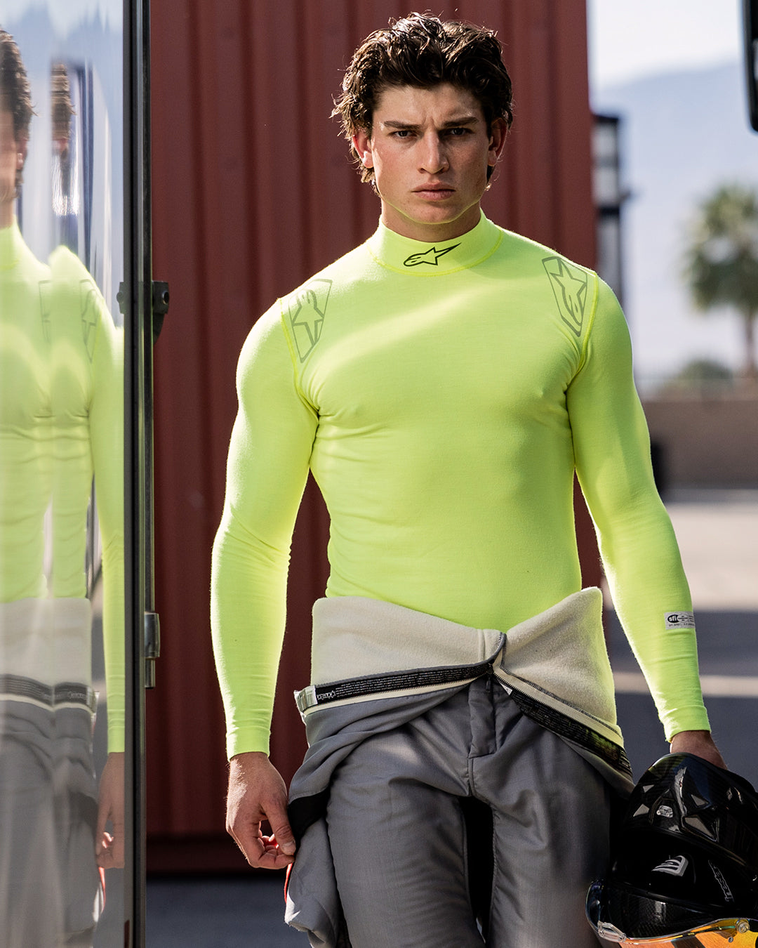 Alpinestars ZX Evo V3 Top - Manga Larga, capa base para automovilismo, amarillo flúor, usada por un piloto masculino con un traje de carreras gris atado a la cintura, sosteniendo un casco negro, con el logo de Alpinestars en el cuello y los hombros, prenda técnica transpirable para automovilismo