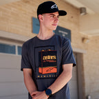 Alpinestars Galun CSF Tee, camiseta de manga corta, gris carbón, gráfico frontal de lata de gasolina vintage con marca 'astars' y números romanos 'MCMLXIII', usada por un hombre con gorra negra con logo, estilo de vida casual de motor