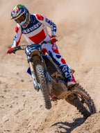 Alpinestars Techstar Fortitude Edición Limitada, pantalones de motocross, en rojo brillante, blanco y azul, usados por un piloto en acción en una pista de tierra, con jersey y botas a juego, equipamiento de carreras off-road de alto rendimiento.