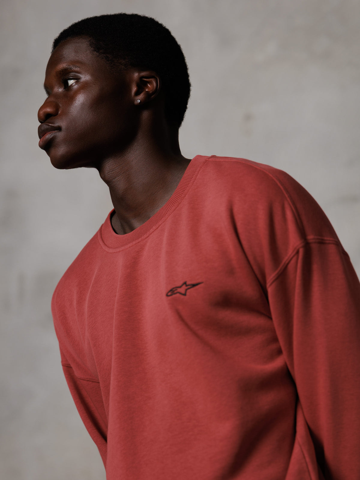 Alpinestars Essentials Ageless Crew, sudadera de cuello redondo en color rojo ladrillo, usada por un modelo masculino de perfil lateral, pequeño logotipo de estrella negro en el pecho, ajuste relajado, forro polar premium de estilo urbano casual