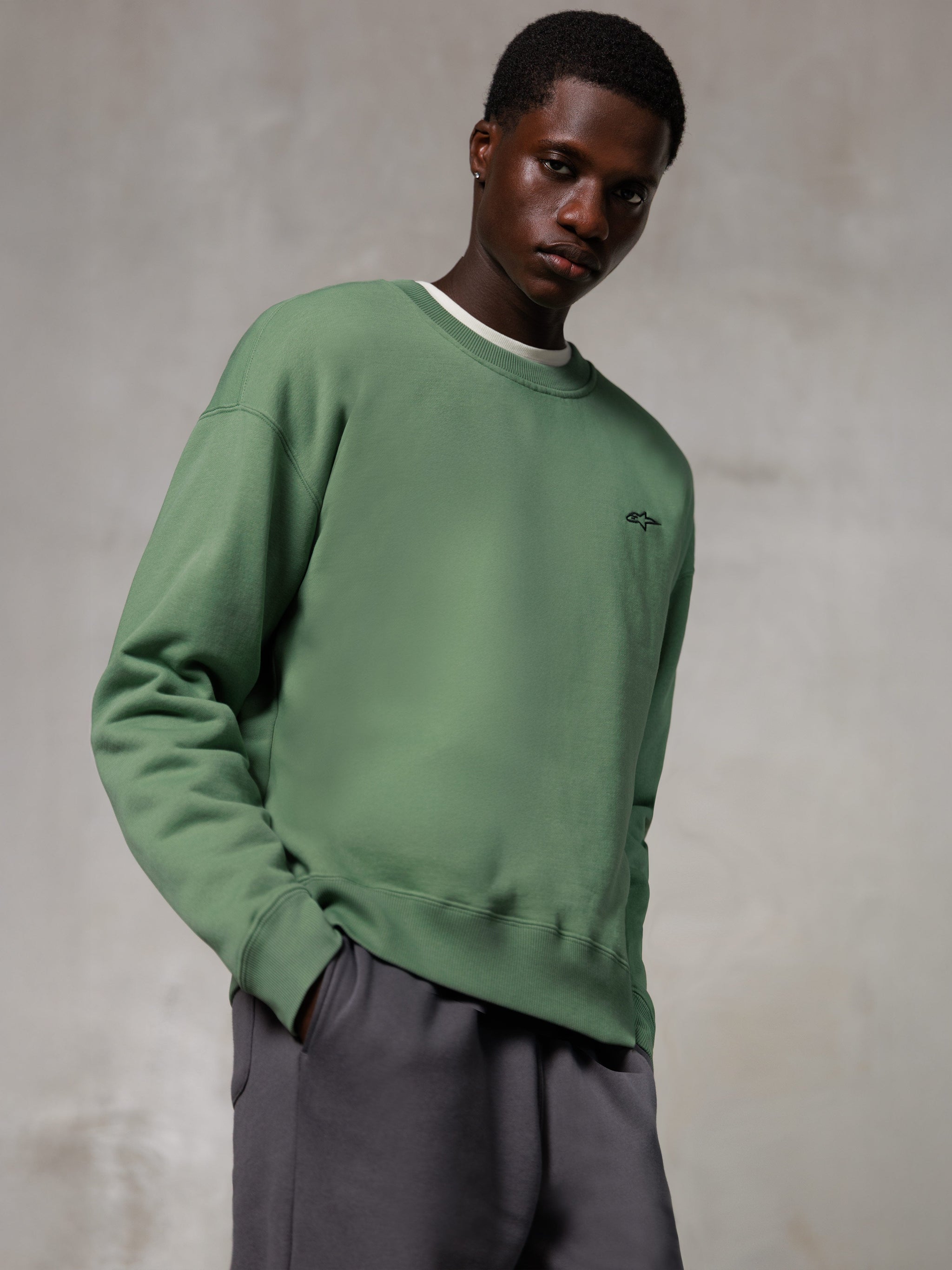 Alpinestars Essentials Ageless Crew, sudadera de cuello redondo, verde cactus, con un pequeño logotipo negro de Alpinestars en el pecho, lucida por un modelo con pantalones grises, ropa urbana de estilo de vida casual