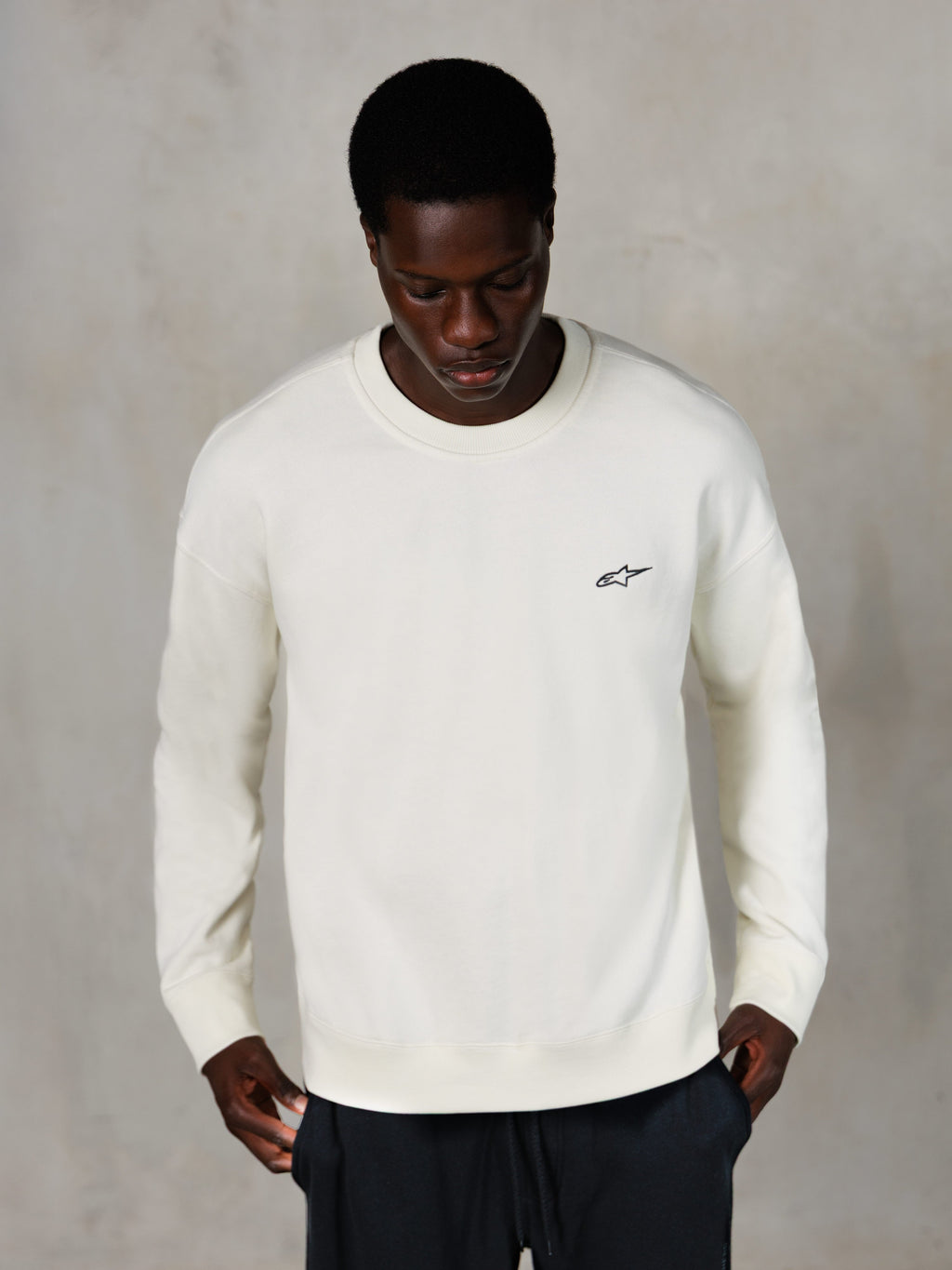 Alpinestars Essentials Ageless Crew, sudadera de cuello redondo, blanco roto, pequeño logo de Alpinestars negro en el pecho, vestida por un modelo, estilo casual lifestyle streetwear, tejido de forro polar suave premium