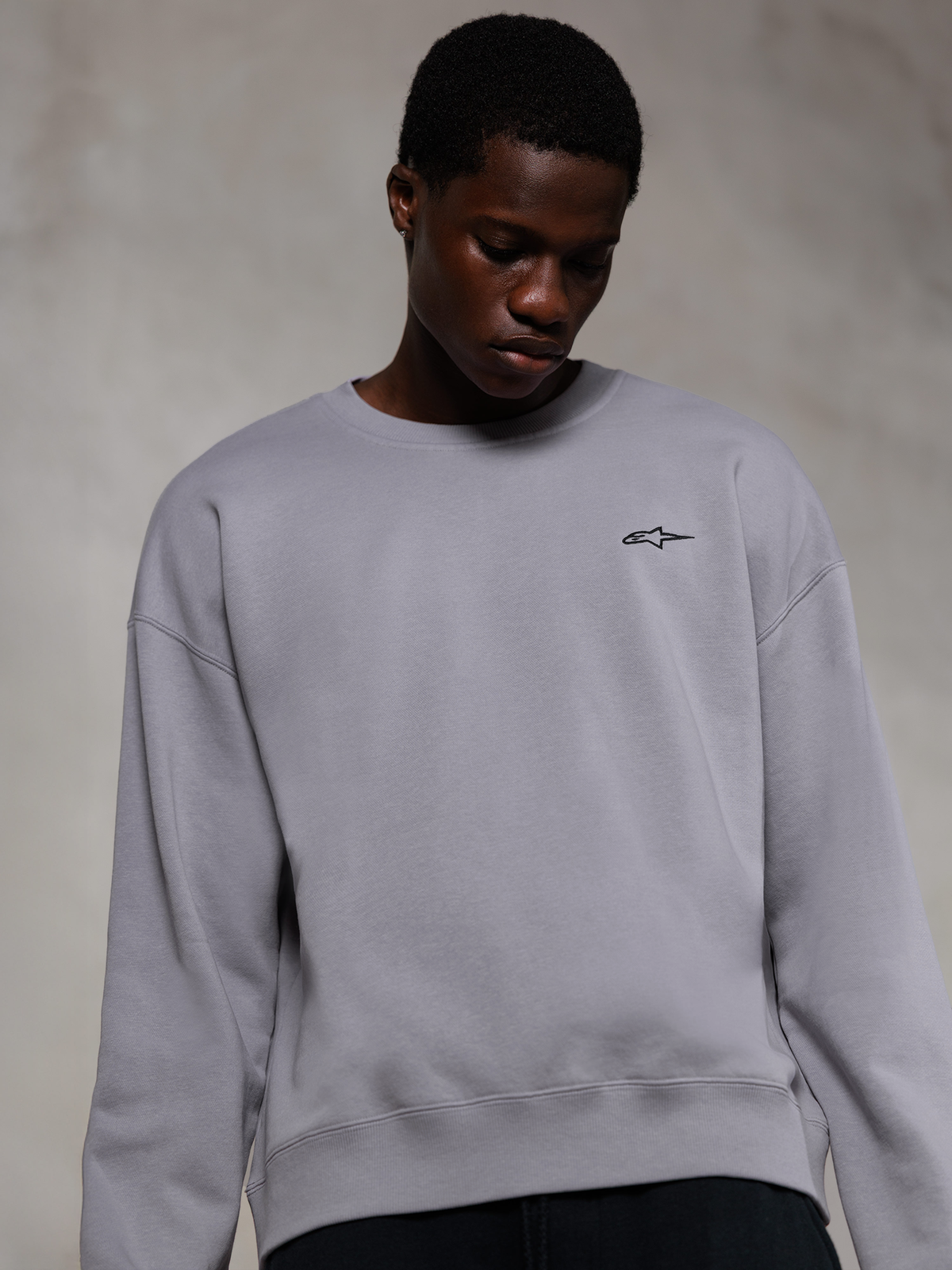 Alpinestars Essentials Ageless Crew, sudadera de cuello redondo premium en color plata, con un pequeño logo negro de Alpinestars en el pecho, lucida por un modelo, ropa urbana de estilo casual con suave textura de forro polar.
