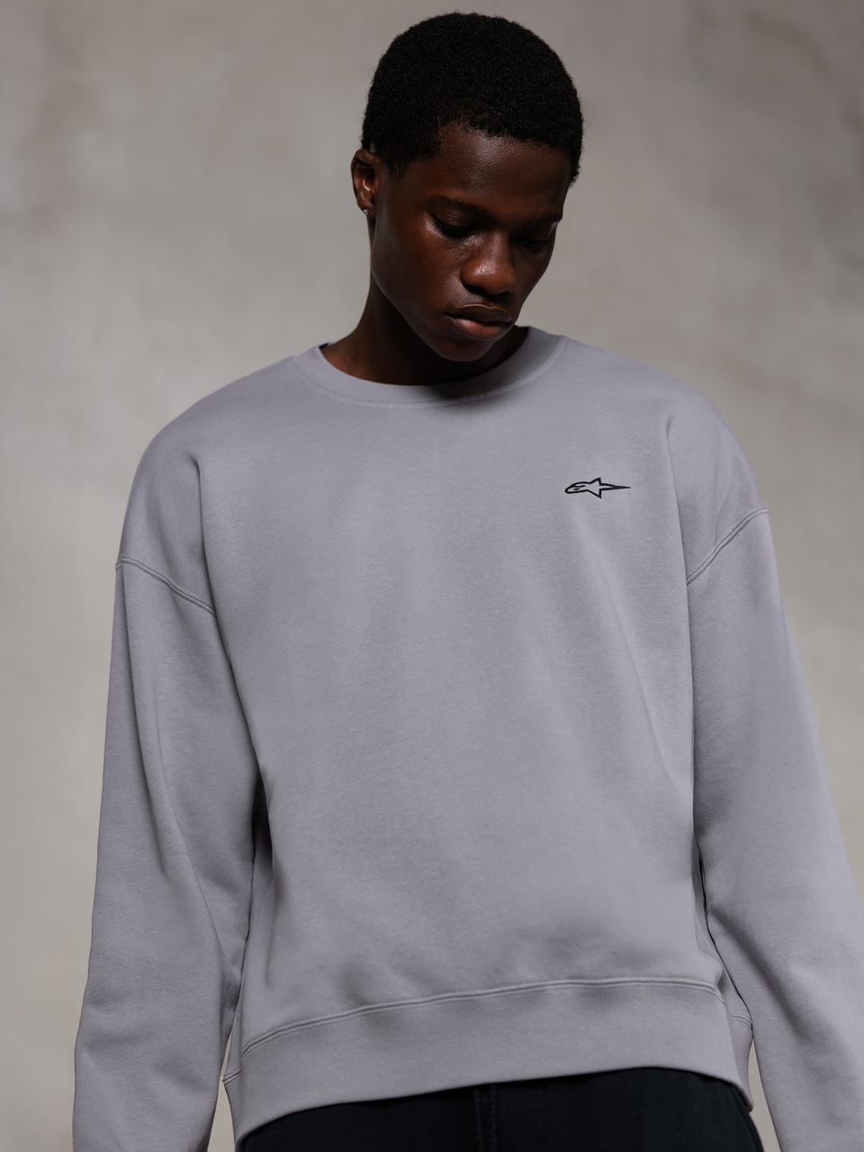 Alpinestars Essentials Ageless Crew, sudadera de cuello redondo premium en color plata, con un pequeño logo negro de Alpinestars en el pecho, lucida por un modelo, ropa urbana de estilo casual con suave textura de forro polar.