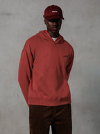 Sudadera con capucha Alpinestars Essentials Ageless, ropa urbana casual, rojo ladrillo, pequeño logotipo negro en el pecho, bolsillo de canguro delantero, combinada con una gorra ASTARS roja a juego y pantalones de pana marrones, diseño minimalista y limpio