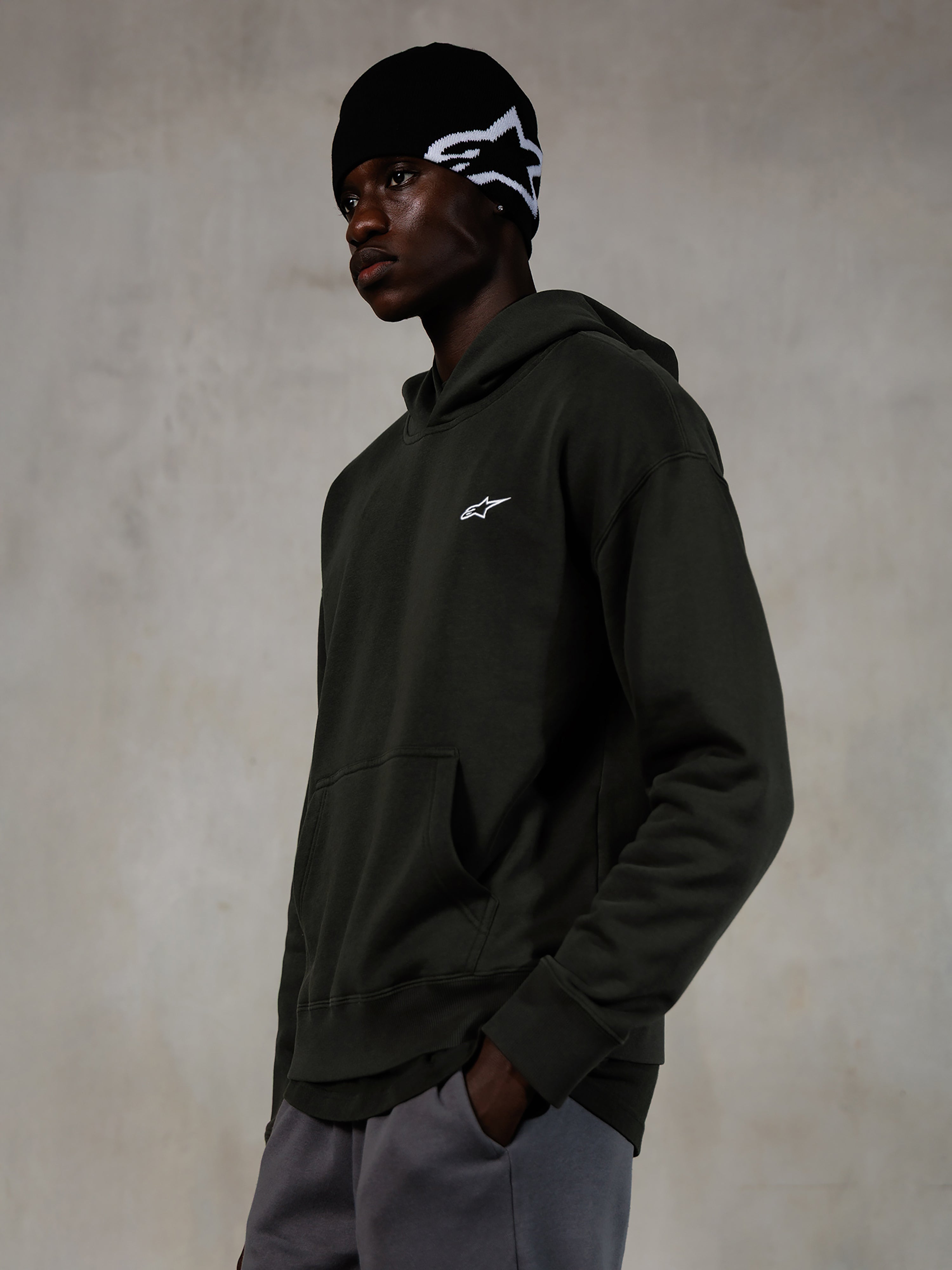 Sudadera con capucha Alpinestars Essentials Ageless, Off Black, modelo masculino luciendo ropa urbana casual, forro polar gris carbón lavado a la piedra con un pequeño logo de estrella blanco, combinado con un gorro beanie negro de Alpinestars y pantalones grises