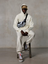 Sudadera con capucha Alpinestars Essentials Ageless, blanco roto, lucida por un modelo sentado en un taburete con pantalones de chándal de forro polar a juego, gorra ASTARS color canela y bandolera, estilo urbano casual inspirado en el automovilismo