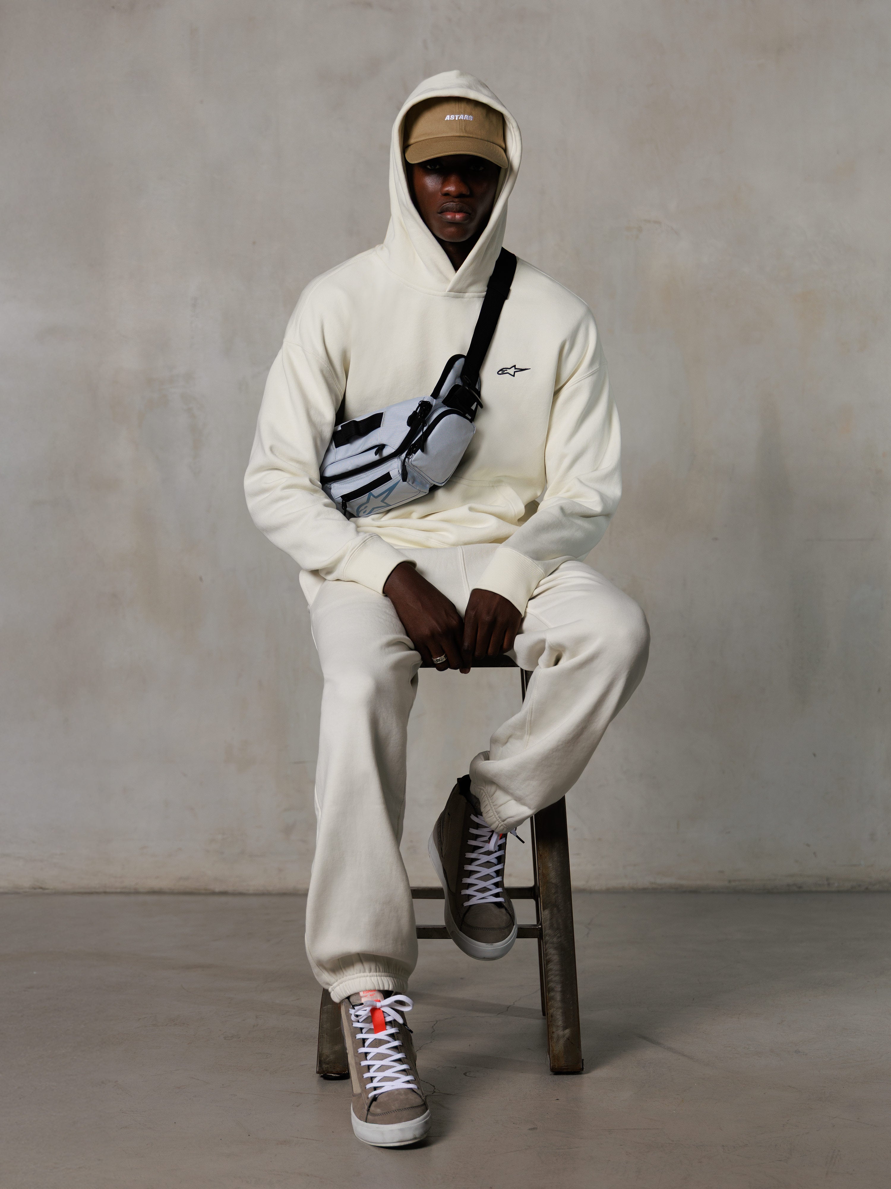 Sudadera con capucha Alpinestars Essentials Ageless, blanco roto, lucida por un modelo sentado en un taburete con pantalones de chándal de forro polar a juego, gorra ASTARS color canela y bandolera, estilo urbano casual inspirado en el automovilismo