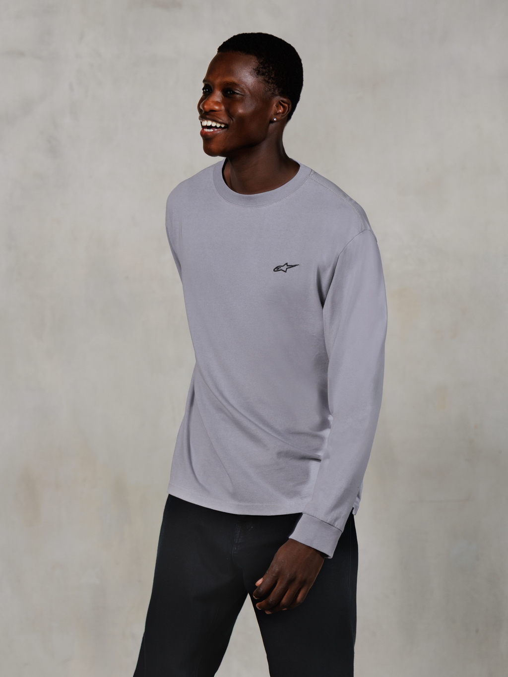 Alpinestars Essentials Ageless LS Tee, Long Sleeve T-Shirt, Silver, small black logo on the chest, worn by a smiling model with dark pants, relaxed contemporary fit, casual motorsport lifestyle apparel