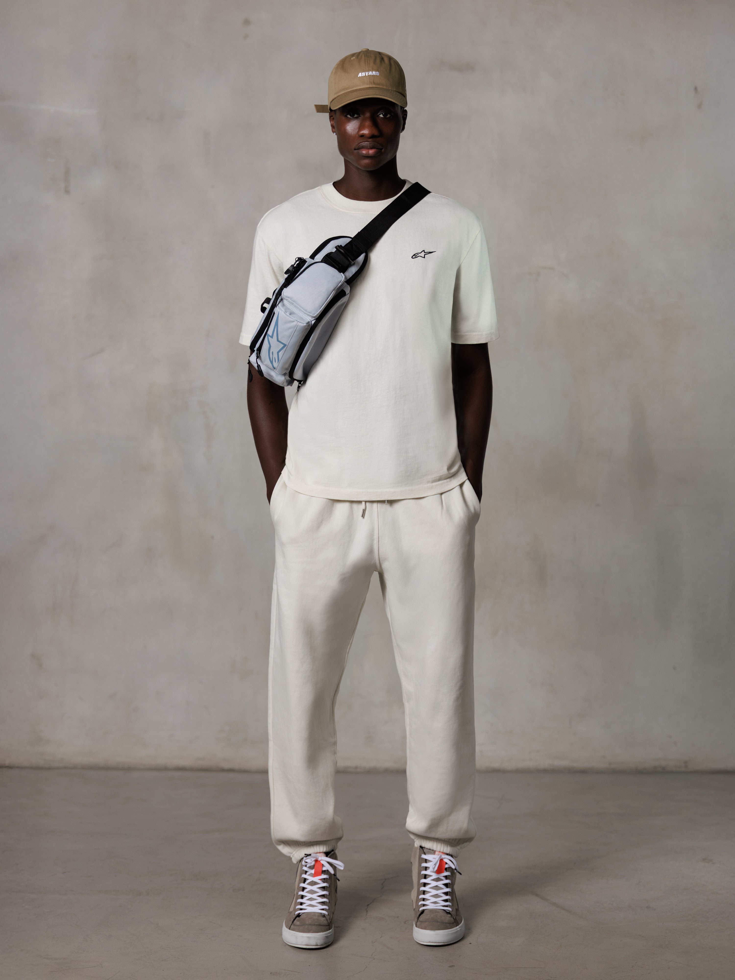 Alpinestars Essentials Ageless SS Tee, camiseta de manga corta, blanco roto, con un pequeño logotipo negro en el pecho, combinada con joggers a juego, gorra de béisbol en color tostado y bolso bandolera gris, estilo urbano casual