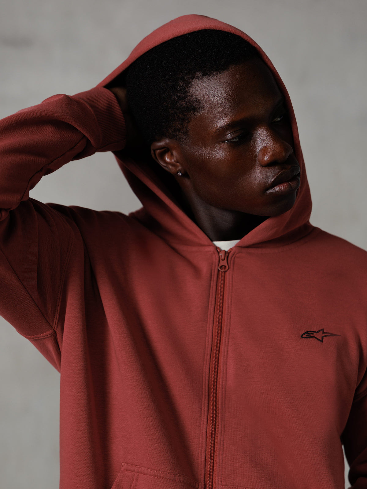 Sudadera con capucha y cremallera Alpinestars Essentials Ageless, color rojo ladrillo, ropa urbana casual, con la capucha puesta y la mano detrás de la cabeza, pequeño logotipo negro de Astars en el pecho, sudadera de forro polar de ajuste relajado