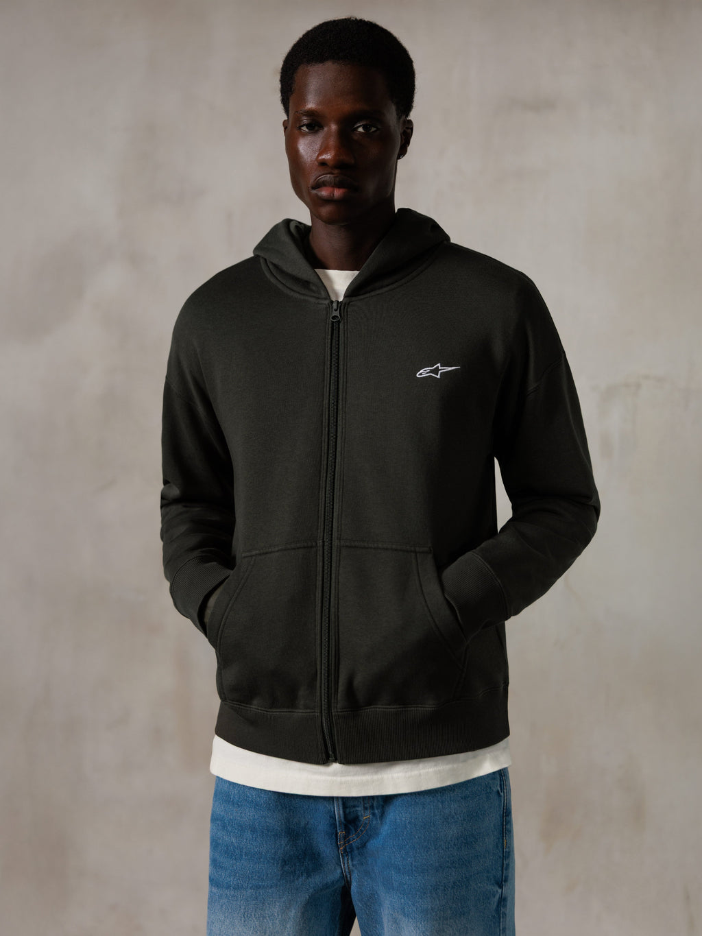 Sudadera con capucha y cremallera Alpinestars Essentials Ageless, Off Black, sudadera con cremallera completa y un pequeño logo blanco de Alpinestars en el pecho, ajuste relajado, llevada por un modelo con vaqueros azules y una camiseta blanca, ropa urbana informal inspirada en el motociclismo