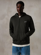 Sudadera con capucha y cremallera Alpinestars Essentials Ageless, Off Black, sudadera con cremallera completa y un pequeño logo blanco de Alpinestars en el pecho, ajuste relajado, llevada por un modelo con vaqueros azules y una camiseta blanca, ropa urbana informal inspirada en el motociclismo