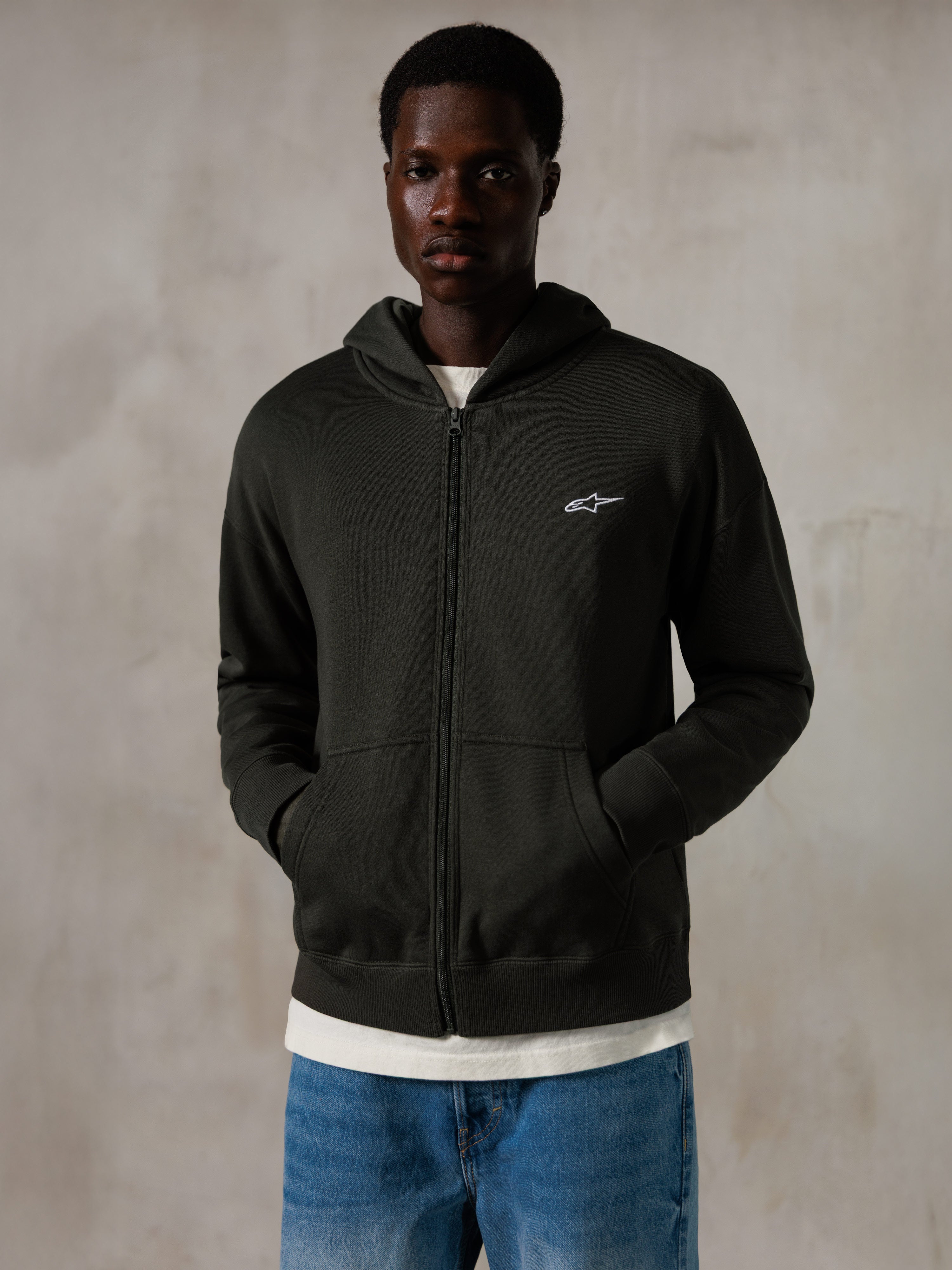 Sudadera con capucha y cremallera Alpinestars Essentials Ageless, Off Black, sudadera con cremallera completa y un pequeño logo blanco de Alpinestars en el pecho, ajuste relajado, llevada por un modelo con vaqueros azules y una camiseta blanca, ropa urbana informal inspirada en el motociclismo