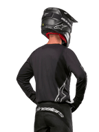 Alpinestars 2024 Fluid Graphite Gear Set, jersey y pantalones de motocross, grafito y plateado, vista trasera con logotipos plateados en la parte baja de la espalda y la cintura, usado con casco negro efecto carbono, diseño de tejido de poliéster ligero para conducción off-road.
