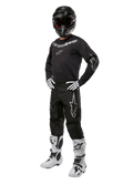 Alpinestars 2024 Fluid Graphite Gear Set, equipo completo de carreras de motocross, negro con logotipos grises y blancos, con jersey y pantalones usados con casco negro a juego, guantes y botas blancas y negras, diseñado para protección todoterreno