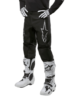 Alpinestars 2024 Fluid Graphite Pants, pantalones de motocross, negros y plateados, con un logotipo plateado de Alpinestars en el muslo, mostrados con botas y guantes de motocross blancos, diseñados para carreras de motos fuera de carretera