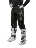 Alpinestars 2024 Fluid Graphite Pants, pantalones de motocross, gris y negro, usados con botas y guantes de motocross en blanco y negro, con un gran logotipo plateado de Alpinestars en el muslo, ajuste deportivo atlético para conducción todoterreno