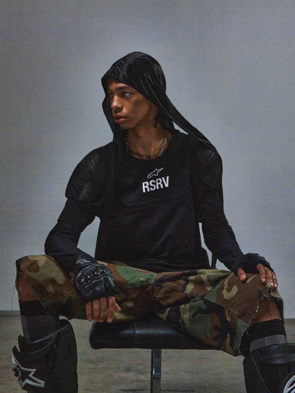 Alpinestars RSRV Nucleon SS Tee, camiseta negra con logo RSRV blanco, estilo urbano táctico, persona sentada con sudadera con capucha negra, pantalones de camuflaje verde y marrón, guantes sin dedos negros, rodilleras negras con logo blanco