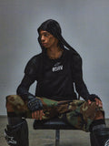 Alpinestars RSRV Nucleon SS Tee, camiseta negra con logo RSRV blanco, estilo urbano táctico, persona sentada con sudadera con capucha negra, pantalones de camuflaje verde y marrón, guantes sin dedos negros, rodilleras negras con logo blanco