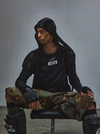 Alpinestars RSRV Nucleon SS Tee, camiseta negra con logo RSRV blanco, estilo urbano táctico, persona sentada con sudadera con capucha negra, pantalones de camuflaje verde y marrón, guantes sin dedos negros, rodilleras negras con logo blanco