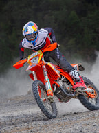Piloto de motocross en acción sobre una moto KTM naranja, vistiendo equipamiento Alpinestars Pro-Dura que incluye un jersey rojo, blanco y negro, pantalones gris oscuro, botas rojas y blancas, y un casco de carreras Red Bull, realizando un giro cerrado en un sendero de grava