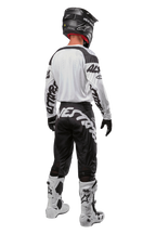 Alpinestars 2024 Racer Hana Pants, pantalones de motocross negros y blancos, vista trasera mostrando el diseño con logo Alpinestars, equipamiento completo con jersey blanco, casco negro y botas blancas protectoras