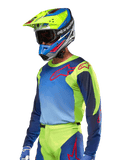 Jersey Alpinestars Racer Hoen 2024, camiseta de motocross, en color amarillo flúor y azul Night Navy con logos rojos, combinada con un casco a juego en amarillo, azul y rojo y guantes azules; equipamiento vibrante para carreras off-road