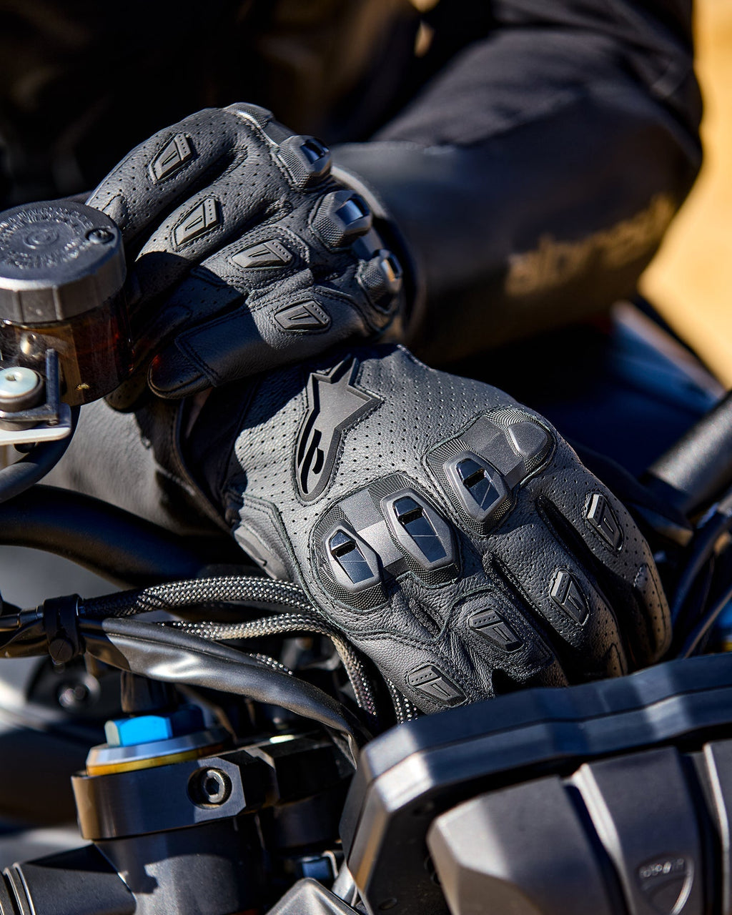 Guantes Alpinestars SP X 7, guantes de moto de cuero perforado negro con protectores de nudillos de TPU y deslizadores en los dedos, mostrados en las manos de un piloto sujetando el manillar de una motocicleta.