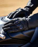 Guantes Alpinestars SP X 7, negros, guantes de moto de puño corto con cuero perforado, manos apoyadas sobre un depósito de combustible oscuro, con protección de nudillos de TPU de doble densidad visible y marca en relieve, diseñados para conducción deportiva y de carretera.