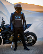 Chaqueta Alpinestars T-Stunt Air, chaqueta textil de corte deportivo, negro, azul oscuro y azul brillante, usada por un piloto de pie junto a una motocicleta Ducati, con un casco de fibra de carbono y equipamiento de moto negro, ambientada en un paisaje desértico al atardecer