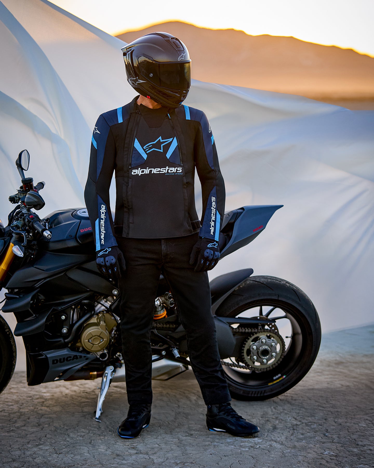 Chaqueta Alpinestars T-Stunt Air, chaqueta textil de corte deportivo, negro, azul oscuro y azul brillante, usada por un piloto de pie junto a una motocicleta Ducati, con un casco de fibra de carbono y equipamiento de moto negro, ambientada en un paisaje desértico al atardecer