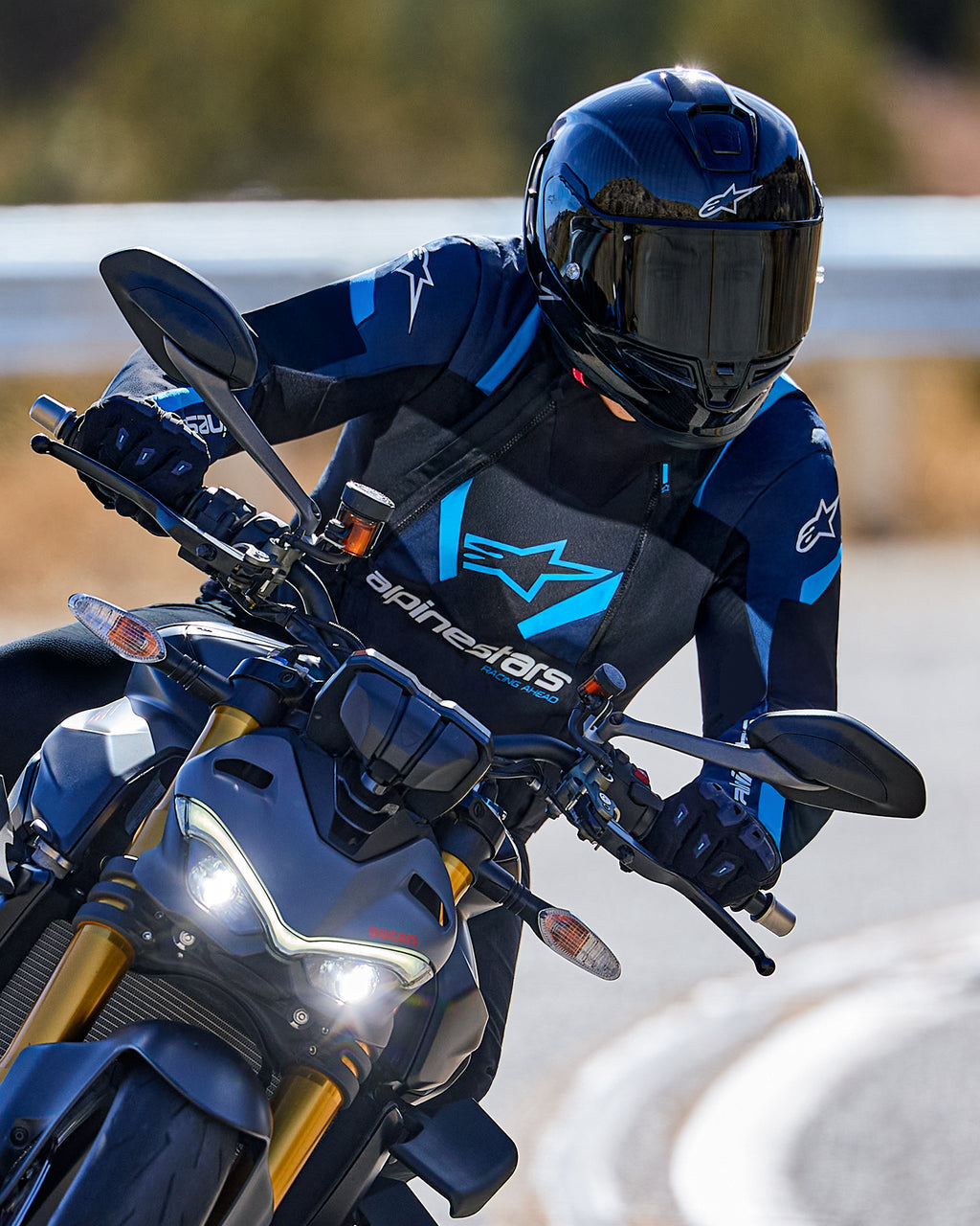 Motociclista inclinándose en una curva usando la chaqueta Alpinestars T-Stunt Air en negro, azul oscuro y azul brillante, con casco y guantes negros, toma de acción en carretera asfaltada, equipo de conducción deportiva de alto rendimiento