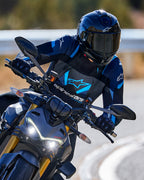 Motociclista inclinándose en una curva usando la chaqueta Alpinestars T-Stunt Air en negro, azul oscuro y azul brillante, con casco y guantes negros, toma de acción en carretera asfaltada, equipo de conducción deportiva de alto rendimiento