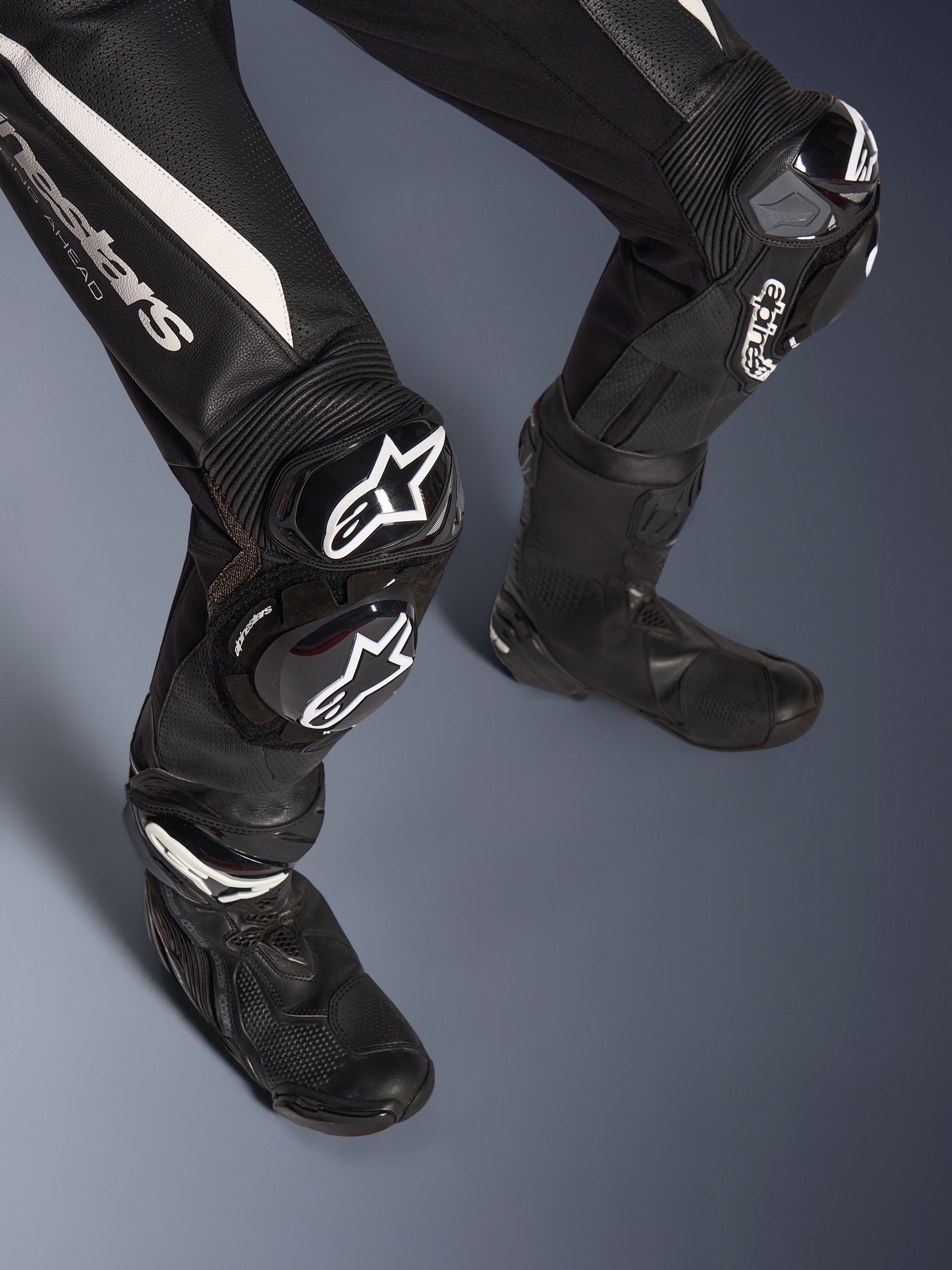 Detalle de las piernas del traje de cuero de 2 piezas Alpinestars Stella GP-R7, negro con detalles blancos, con protecciones de rodilla GP DFS RR y deslizadores con la marca Alpinestars, combinado con botas de competición negras, diseñado para el rendimiento en pista.