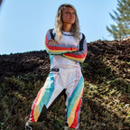 Alpinestars Stella Techstar Pants 2024 para mujer, pantalones de motocross con jersey a juego, diseño Black Multicolor con base blanca y rayas vibrantes en azul, amarillo y rojo, piloto femenina de pie con los brazos cruzados en una pista de tierra, equipamiento profesional de carreras off-road