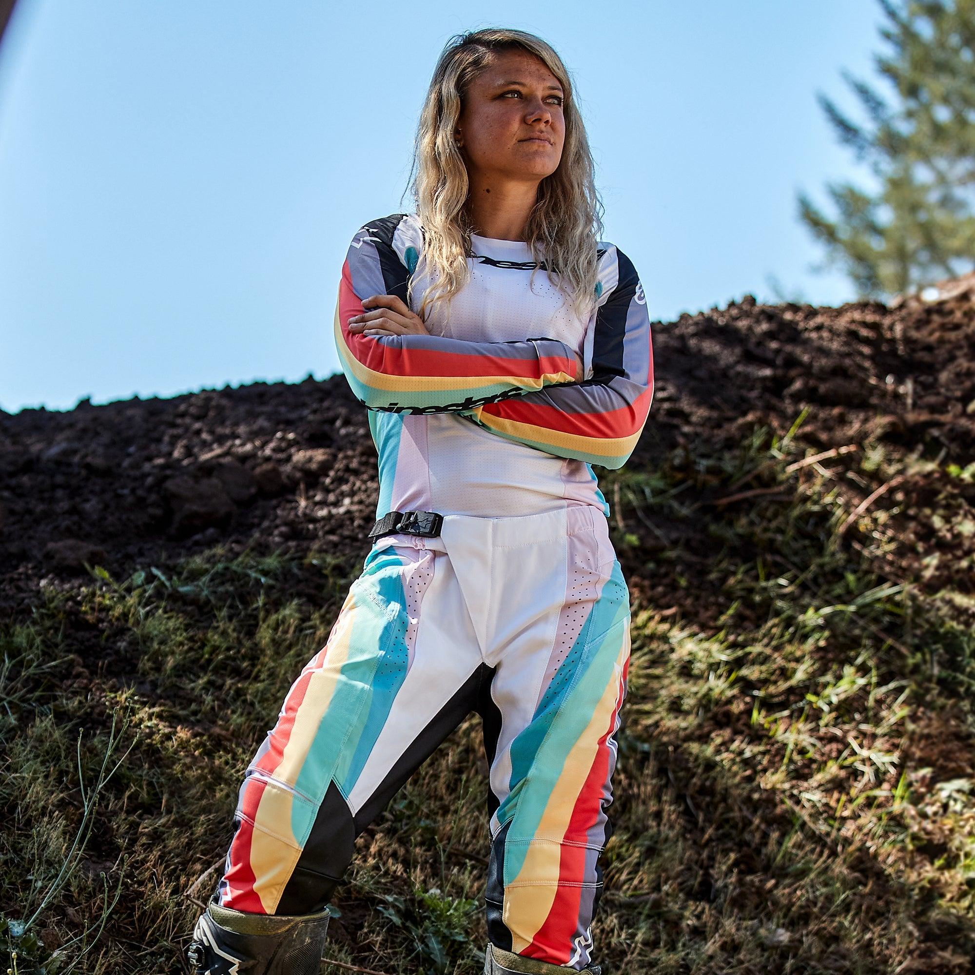 Alpinestars Stella Techstar Pants 2024 para mujer, pantalones de motocross con jersey a juego, diseño Black Multicolor con base blanca y rayas vibrantes en azul, amarillo y rojo, piloto femenina de pie con los brazos cruzados en una pista de tierra, equipamiento profesional de carreras off-road