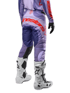 Alpinestars Techstar Ocuri Pants 2024, pantalones de motocross para hombre, color morado con detalles en naranja vibrante y azul oscuro, vista trasera mostrando el logo de Alpinestars, diseño ergonómico con paneles reforzados, usados con botas de motocross blancas.