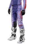 Alpinestars 2024 Techstar Ocuri Pants, pantalones de motocross para hombre, color púrpura con detalles en naranja vibrante y logos rojos, diseño ergonómico con paneles elásticos, combinados con botas blancas de motocross y guantes negros.