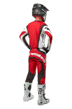 Alpinestars 2024 Techstar Ocuri Pants, pantalones de motocross, Rojo Marte Blanco Negro, vista trasera de un piloto de motocross con equipo completo de competición, incluyendo jersey a juego, casco negro y botas blancas y negras, resaltando la marca Alpinestars y la construcción de tejido duradero.