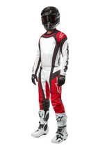 Piloto de motocross con equipamiento completo de Alpinestars, pantalones Techstar Ocuri 2024 en color Mars Red, blanco y negro, jersey a juego, casco negro con gafas, guantes negros y blancos, y botas de motocross blancas; atuendo profesional de carreras todoterreno