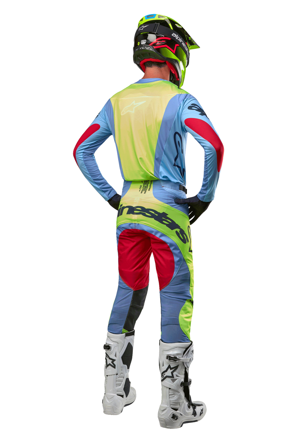 Piloto de motocross visto desde atrás con pantalones Techstar Ocuri 2024 y jersey a juego, azul claro, amarillo flúor y rojo, botas blancas y negras, casco negro con detalles neón, equipamiento coordinado de Alpinestars para carreras todoterreno